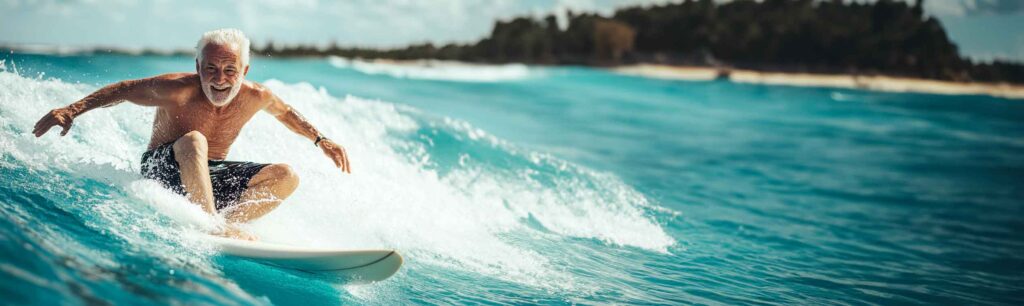 Older gentleman riding a wave on a surfboard smiling in the sun