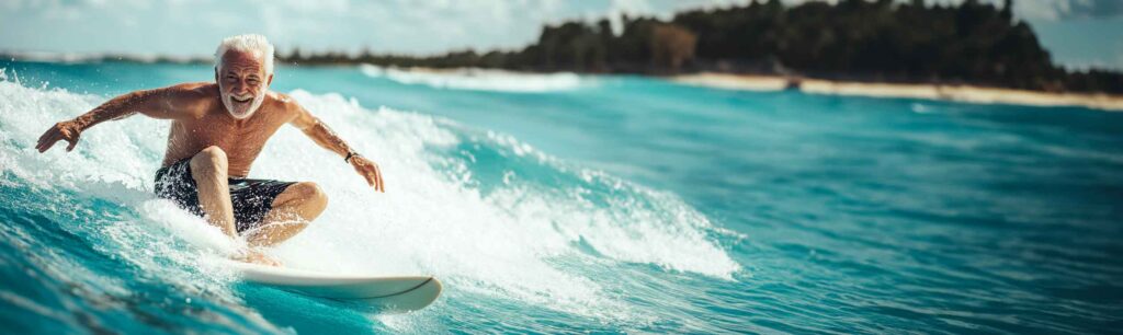 Older gentleman riding a wave on a surfboard smiling in the sun